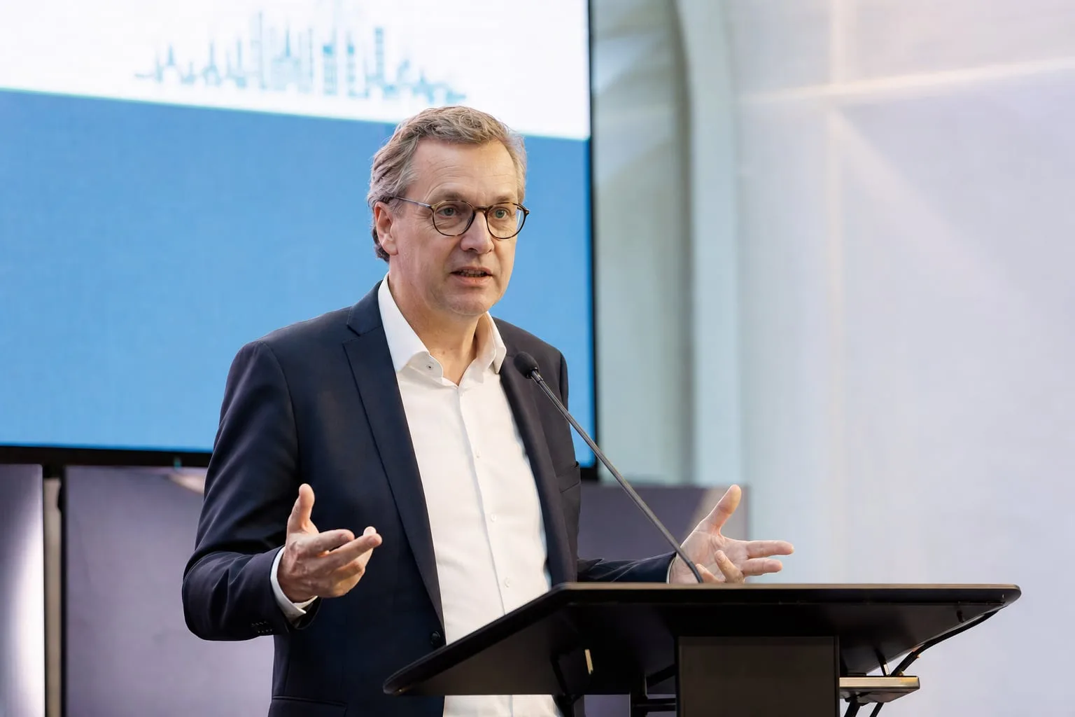 Redner am Rednerpult mit Schwanenhalsmikrofon, hinter ihm die Präsentation auf der Leinwand